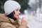 A young girl in a white hat warms her hands. It stands in the city in winter under the snow