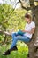 Young girl in white T-shirt is sitting on a tree in the garden and reading book on ebook reader. Great idea and activity to spend