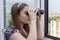 A young girl from the window is watching the street and neighboring houses through binoculars. Concept: spying on neighbors, gathe