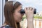 A young girl from the window is watching the street and neighboring houses through binoculars. Concept: spying on neighbors, gathe