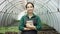 Young girl worker stands near new seedlings with her tablet in a greenhouse and smiles at camera