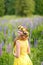 Young girl in yellow dress walking on meadow with lilac lupines. Warm summer day. Flower wreath for head