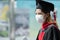 Young graduated Asian woman wear black gown, cap with red tassels and face mask holding diploma after graduation.