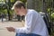 A young guy of 20-25 years old with a backpack sits at the metal fence of the park and looks at the phone against the backdrop of