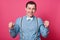 Young guy with blue shirt, suspenders and bow tie, celebrating something with his fists up, isolated on pink background. Handsome