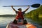A young guy in a campground and glasses sits in a kayak and holds an oar over his head. On the sparing plan, a lake and a