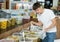 Young guy chooses marinated olives in supermarket, scooping them from bucket with ladle into plastic bag