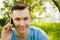 Young guy communicates on a smartphone and sitting on green grass with dandelions on a background of trees