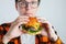 A young guy with glasses holding a fresh Burger. A very hungry student eats fast food. Hot helpful food. The concept of gluttony a