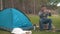 A young guy sits on a large log near the blue tent and makes a photo on the phone.