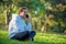 Young guy speaks on mobile phone sitting on the green lawn. Young smiling man is talking on the phone in a park, a sunny summer