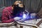 Young guy welder in a checkered red shirt welds a stainless steel pipe for car exhaust using agronomic welding to protect his eyes