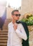 Young guy in a white shirt and sunglasses stands near the stairs and flowers at sunset in the sun`s rays