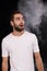 A young guy in a white T-shirt on a black background with smoke looks to the side