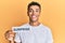 Young handsome african american man holding paper with surprise text over yellow background looking positive and happy standing