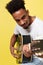 Young handsome African American retro styled guitarist playing acoustic guitar on yellow gold background.