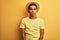 Young handsome arab man wearing t-shirt and summer hat over isolated yelllow background with serious expression on face