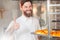 A young handsome baker shows his thumb up with a sheet of fresh croissants in his hands against the backdrop of an oven.