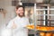 A young handsome baker shows his thumb up with a sheet of fresh croissants in his hands against the backdrop of an oven.