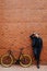 Young handsome bearded business man in suit and glasses with hands on head standing near his bicycle on brick wall background