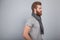 Young handsome bearded man standing by the grey wall rotated in profile wearing scarf and grey t-shirt