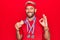 Young handsome blond sportsman with beard winning medals over isolated red background doing ok sign with fingers, smiling friendly