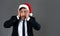 Young handsome caucasian guy in business suit and Santa hats stands on gray background in studio very surprised hands on cheeks