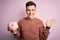 Young handsome caucasian man holding piggy bank for savings over pink background very happy and excited, winner expression