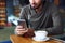 Young handsome cheerful hipster guy at the restaurant using a mobile phone, hands close up. Selective focus.