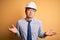 Young handsome chinese architect man wearing safety helmet and tie over yellow background clueless and confused expression with