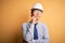 Young handsome chinese architect man wearing safety helmet and tie over yellow background with hand on chin thinking about