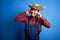 Young handsome chinese farmer man wearing apron and straw hat over blue background covering ears with fingers with annoyed