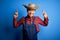 Young handsome chinese farmer man wearing apron and straw hat over blue background gesturing finger crossed smiling with hope and