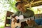 Young handsome and happy mixed ethnicity man in hipster style chilling outdoors playing guitar relaxed at tranquil tropical garden