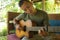 Young handsome and happy mixed ethnicity man in hipster style chilling outdoors playing guitar relaxed at tranquil tropical garden