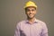 Young handsome Indian businessman wearing hardhat against gray background