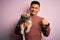 Young handsome latin man holding cute puppy pet over isolated pink background with surprise face pointing finger to himself