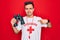 Young handsome lifeguard man wearing t-shirt with red cross and whistle using binoculars with surprise face pointing finger to