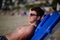 A young handsome man of 25-30 years old, a brunette, in weed sunglasses, lies relaxed on a lounger on a sandy sea beach, takes a