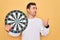 Young handsome man with blue eyes holding dartboard over isolated yellow background pointing and showing with thumb up to the side