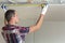 Young handsome man in casual clothing takes measurement of drywall suspended ceiling connected to metal frame on ceiling insulated