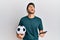 Young handsome man holding football ball looking at smartphone bet app angry and mad screaming frustrated and furious, shouting