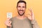 Young handsome man holding reminder with coronavirus message over yellow background doing ok sign with fingers, smiling friendly