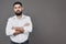 Young handsome man leaning against grey wall with arms crossed. A serious young man with a beard looks at the camera