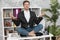 Young handsome man manager sitting in yoga position on the table at his office looking relaxed at the end of working day