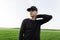 Young handsome man in stylish cap in black trendy shirt posing standing in a field with green grass. Attractive guy model enjoys