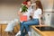 Young handsome man surprising his beloved wife with roses and gift on saint valentine`s day in the kitchen, happy romantic