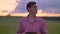 Young handsome man typing on tablet and standing on wheat or rye field, pink sky above with clouds