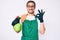 Young handsome man wearing apron holding sprayer doing ok sign with fingers, smiling friendly gesturing excellent symbol