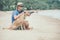 Young handsome man wearing blue t-shirt, hat and sunglasses, sitting on the beach with the dog